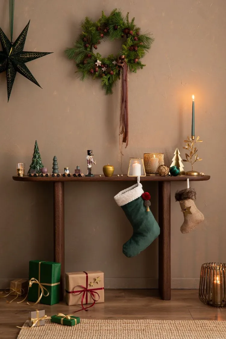 une console en bois est ornée d'une couronne naturelle suspendue au mur, de bougies dorées, et de petites figurines festives comme un casse noisette et des sapins miniatures