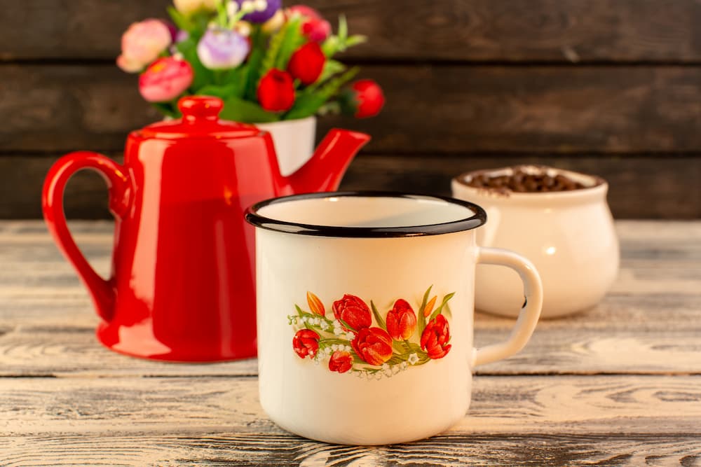 tasse en métal décorée de fleurs devant une théière rouge sur table en bois