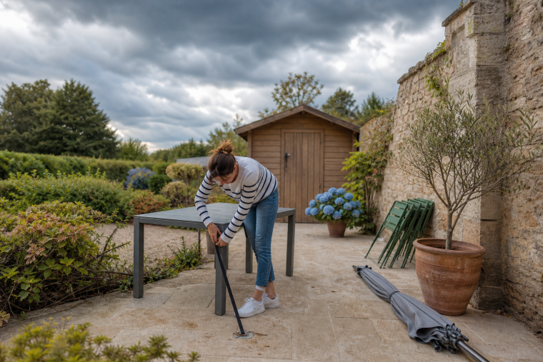 Femme arrimant le mobilier de jardin avant une tempête, parasol couché et pot d'olivier descendu sur une terrasse française