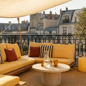 Terrasse parisienne aménagée comme un salon d'hôtel avec canapé bas, coussins jaune beurre et lampe nomade au coucher du soleil