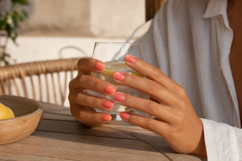 Mains bronzées présentant la manucure aura nails corail terminée en scène de vie estivale