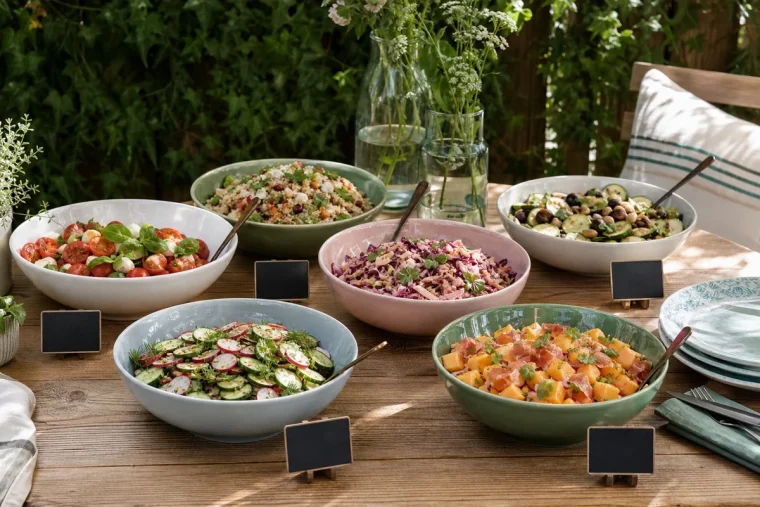 Buffet de salades d'été dressé sur table en bois dans un jardin, six saladiers colorés et étiquettes ardoise