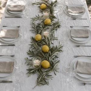 Table d'été dressée avec chemin de table en branches d'olivier fraîches, citrons jaunes et bougies blanches sur nappe en lin