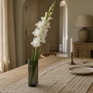 Une tige de glaïeul blanc dans un vase cylindrique en verre fumé, sur un chemin en lin beige et une table en chêne clair